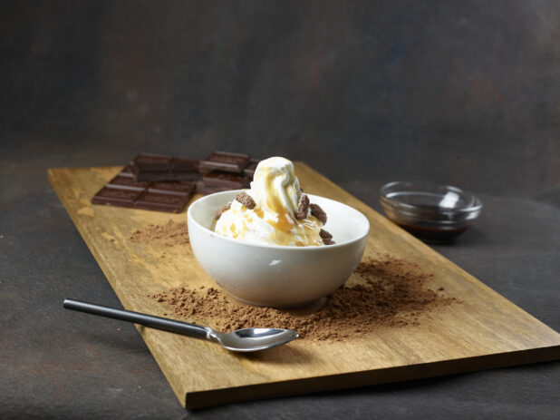 Bowl of vanilla ice cream topped with chocolate cookie bits and caramel in a white bowl on a wooden board