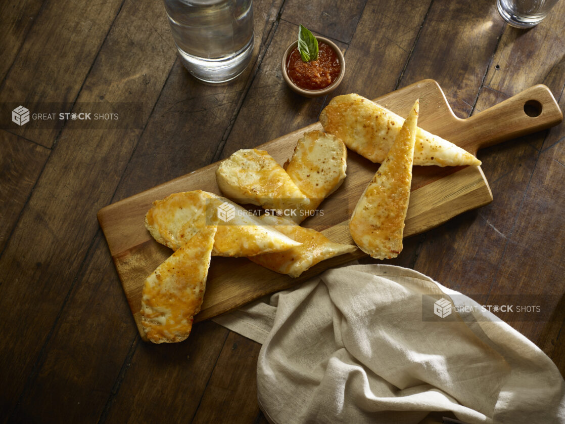 Slices of garlic bread with cheese on a cutting board with dipping sauce on a wooden table