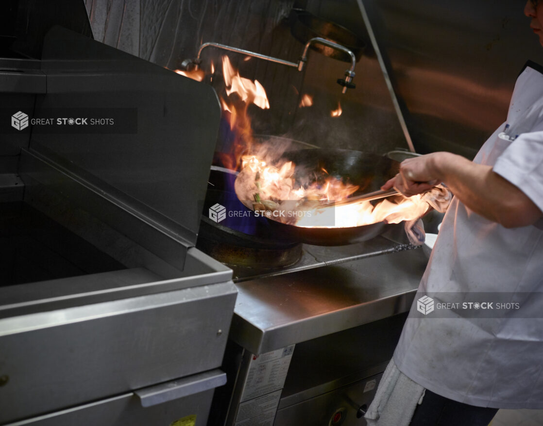 Action shot of a chef in a white apron stirring the contents of a flaming wok