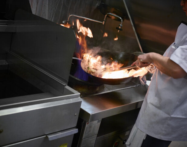 Action shot of a chef in a white apron stirring the contents of a flaming wok