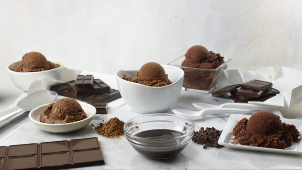 Many bowls of chocolate ice cream surrounded by bars of chocolate, cocoa, chocolate chips and spoons