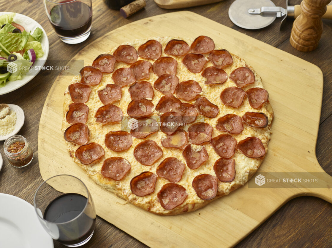 Whole pepperoni pizza on a wooden paddle with red wine and a side salad