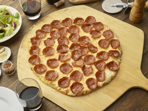 Whole pepperoni pizza on a wooden paddle with red wine and a side salad