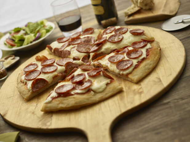 Sliced pepperoni pizza on a wooden paddle with garden salad and a drink