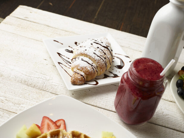 Chocolate drizzled croissant with a berry shake on a light wooden background