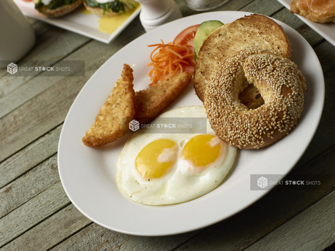 Breakfast plate of two eggs, sesame seed bagel and hash browns on a wooden background