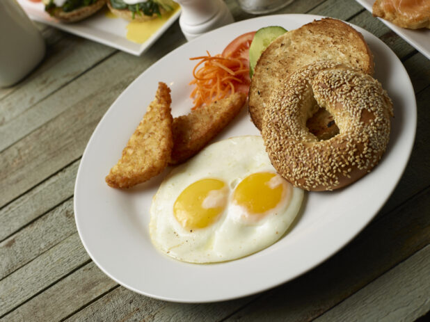Breakfast plate of two eggs, sesame seed bagel and hash browns on a wooden background