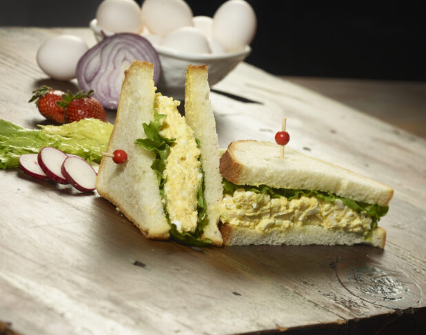 Egg salad sandwich cut in half on a wooden table with a bowl of eggs and other ingredients in the background