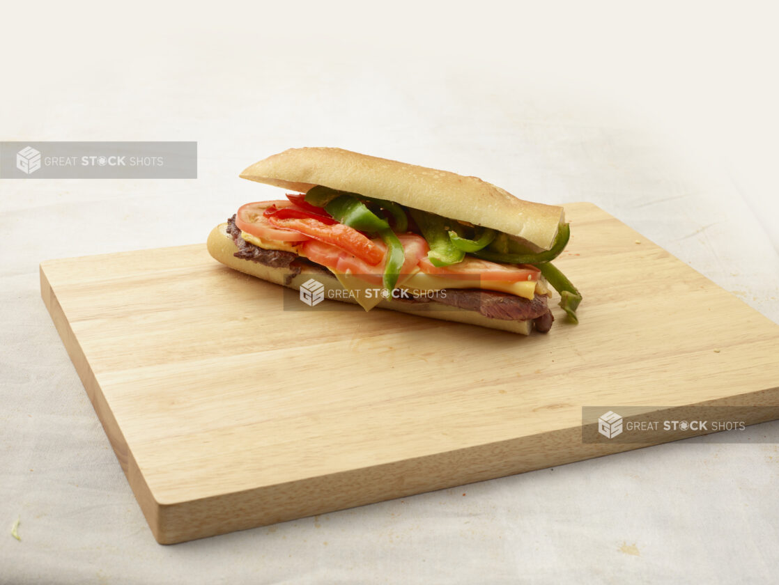 Steak and cheese sandwich with vegetables sitting on top of a wooden cutting board