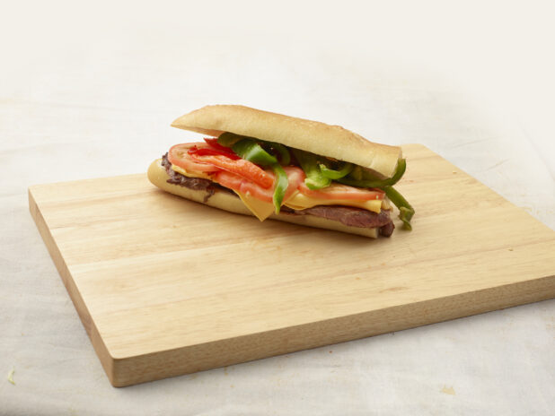 Steak and cheese sandwich with vegetables sitting on top of a wooden cutting board