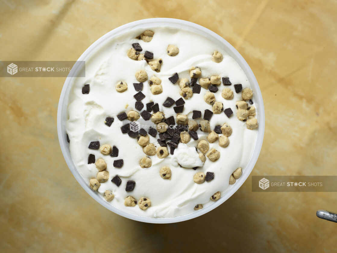 Overhead view of a large tub of vanilla ice cream with chocolate chips and cookie dough on a yellow background