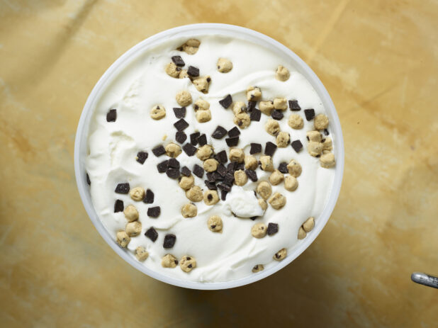 Overhead view of a large tub of vanilla ice cream with chocolate chips and cookie dough on a yellow background