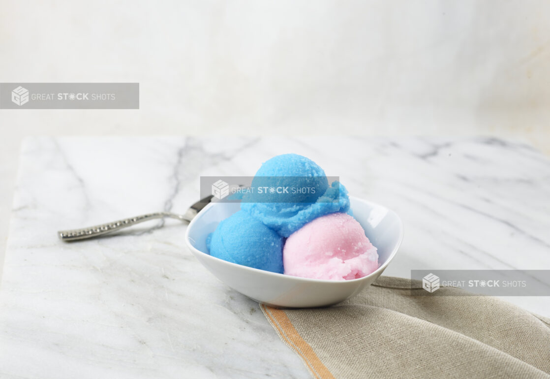 Scoops of blue and pink gelato in a white bowl on a marbe background