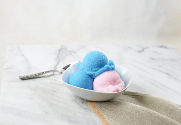 Scoops of blue and pink gelato in a white bowl on a marbe background