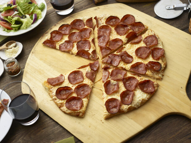 Sliced pepperoni pizza on a peel with a side salad and glasses of red wine