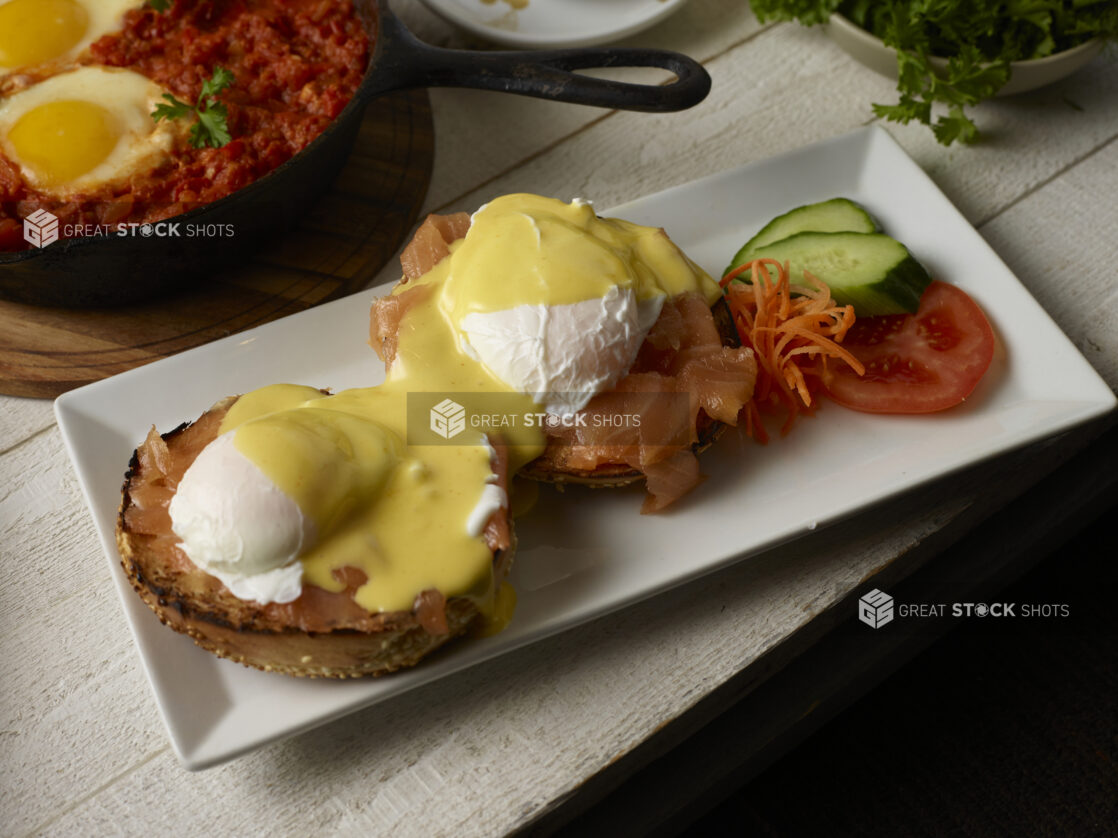 Eggs benedict with salmon (Eggs Royal) on a toasted sesame bagel with a shakshuka skillet in the background