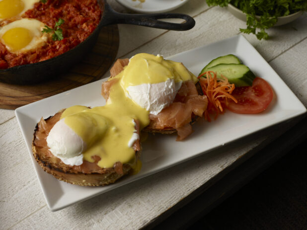 Eggs benedict with salmon (Eggs Royal) on a toasted sesame bagel with a shakshuka skillet in the background