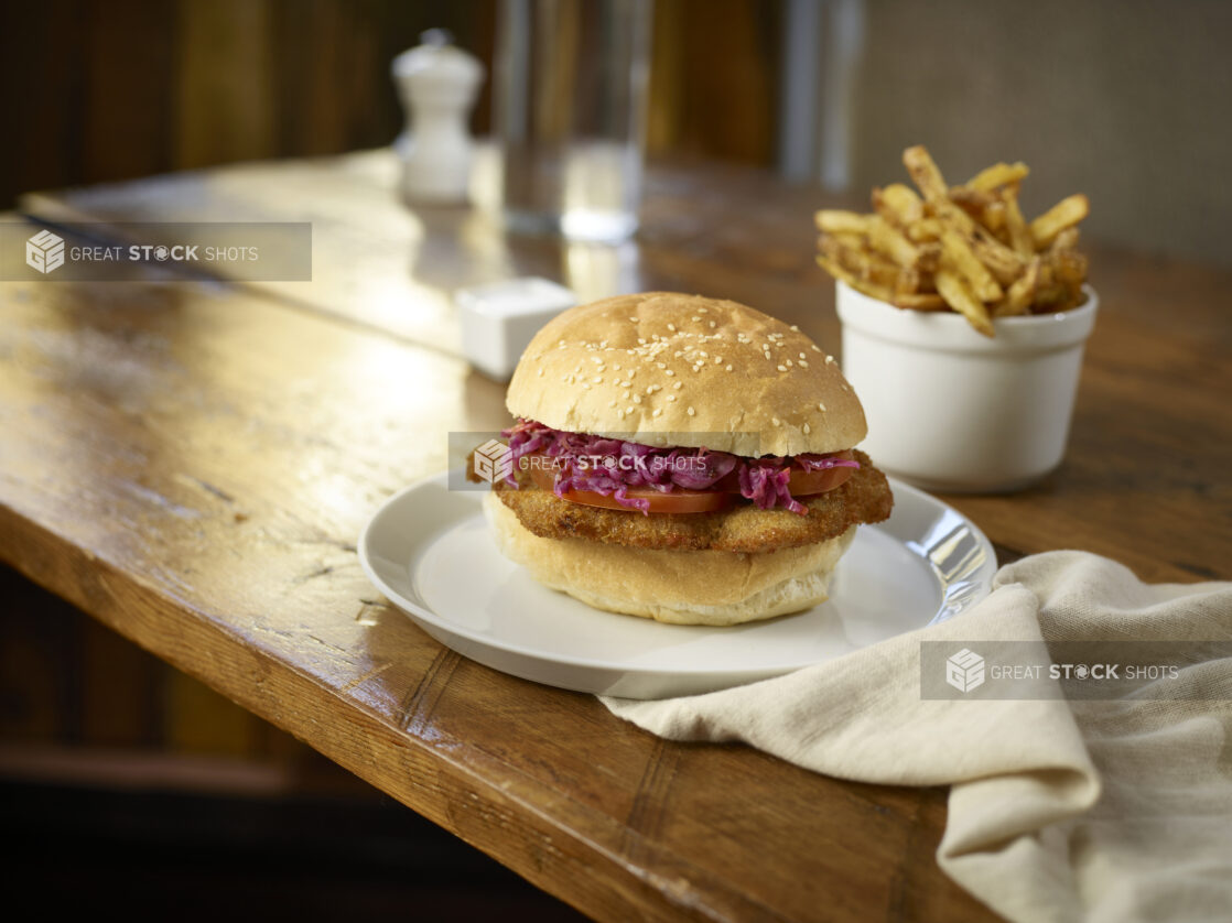 Fried chicken sandwich with coleslaw on a bun with a side of fries, white dishes, wood tabletop