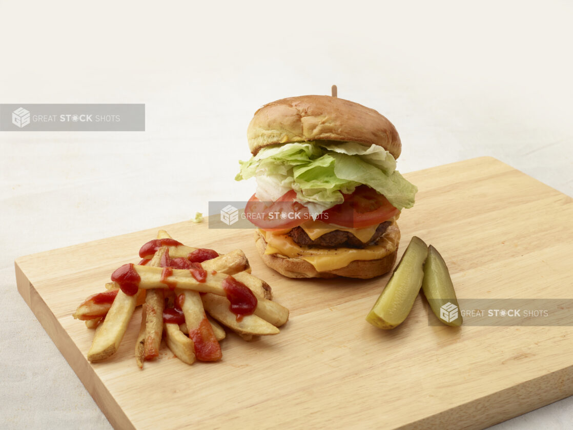 Hamburger with cheese, lettuce and tomato on a bun with french fries and dill pickle spears on a wooden board