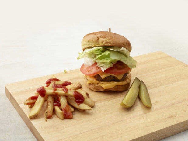 Hamburger with cheese, lettuce and tomato on a bun with french fries and dill pickle spears on a wooden board