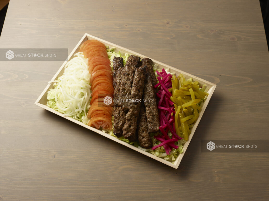 Overhead view of a wooden platter with sliced, onion, tomatoes, turnip, pickles and beef kebab on a wooden background