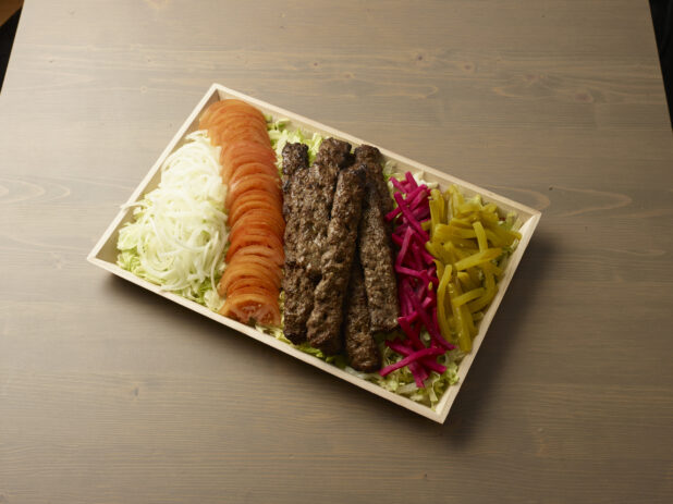 Overhead view of a wooden platter with sliced, onion, tomatoes, turnip, pickles and beef kebab on a wooden background