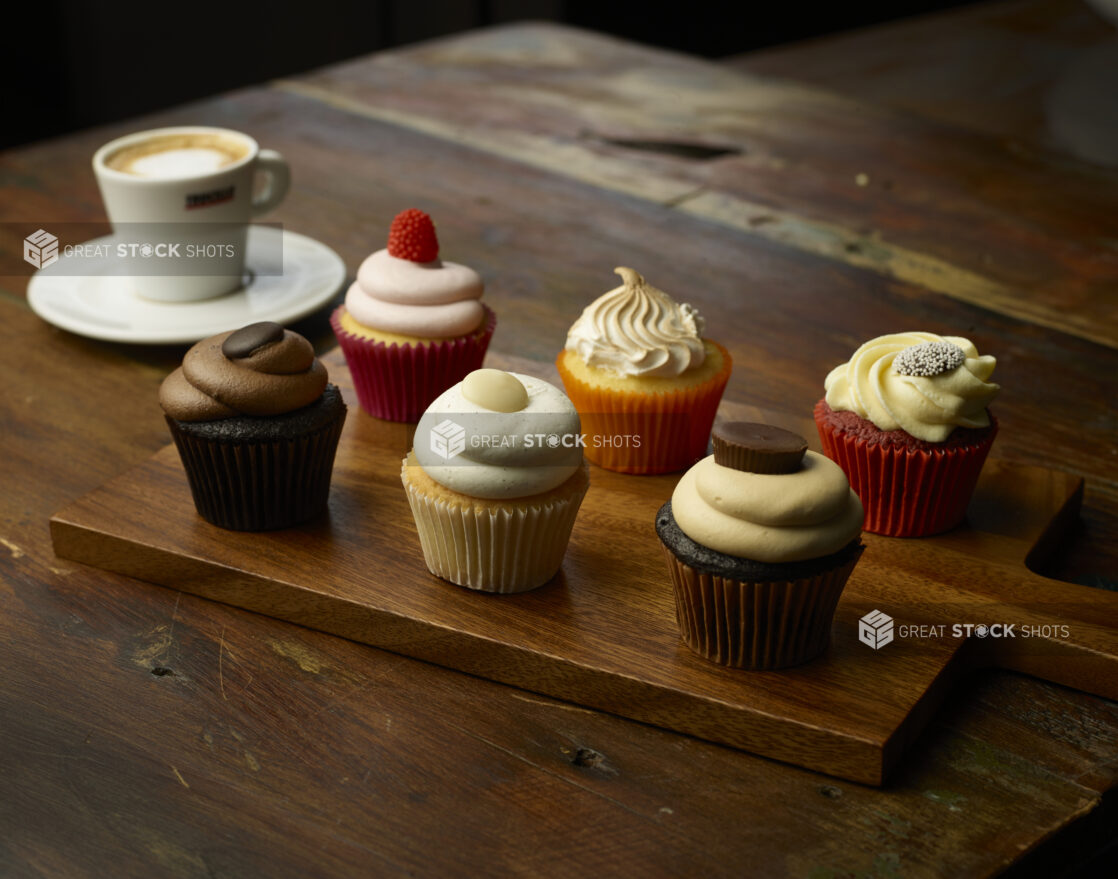 Half dozen different flavored cupcakes on wooden cutting board with espresso