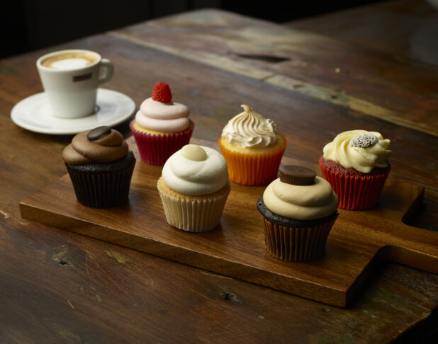 Half dozen different flavored cupcakes on wooden cutting board with espresso
