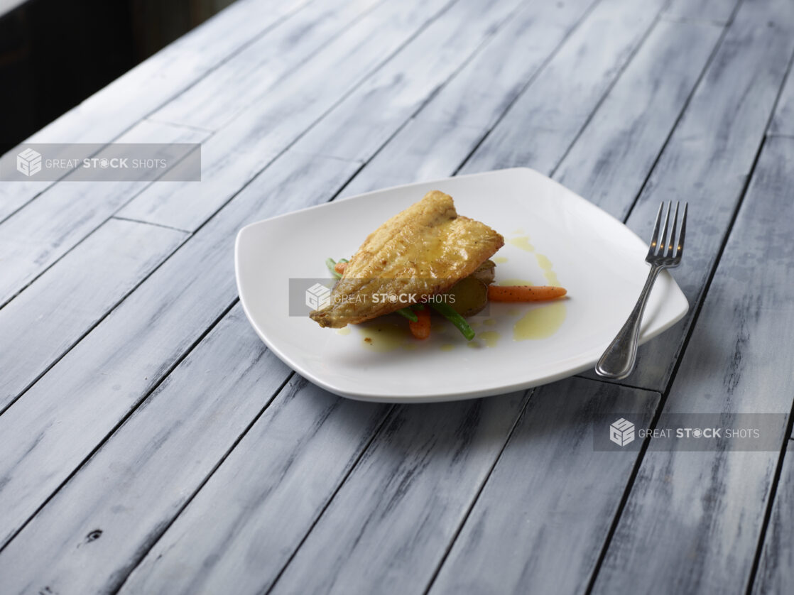 Pan fried white fish on top of roasted vegetables with an oil drizzle on a white plate on a wooden background