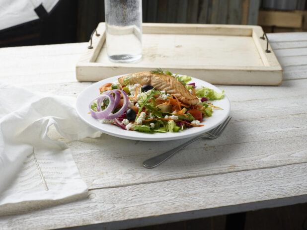 Greek salad with grilled salmon in a white bowl on a whitewashed wooden background,