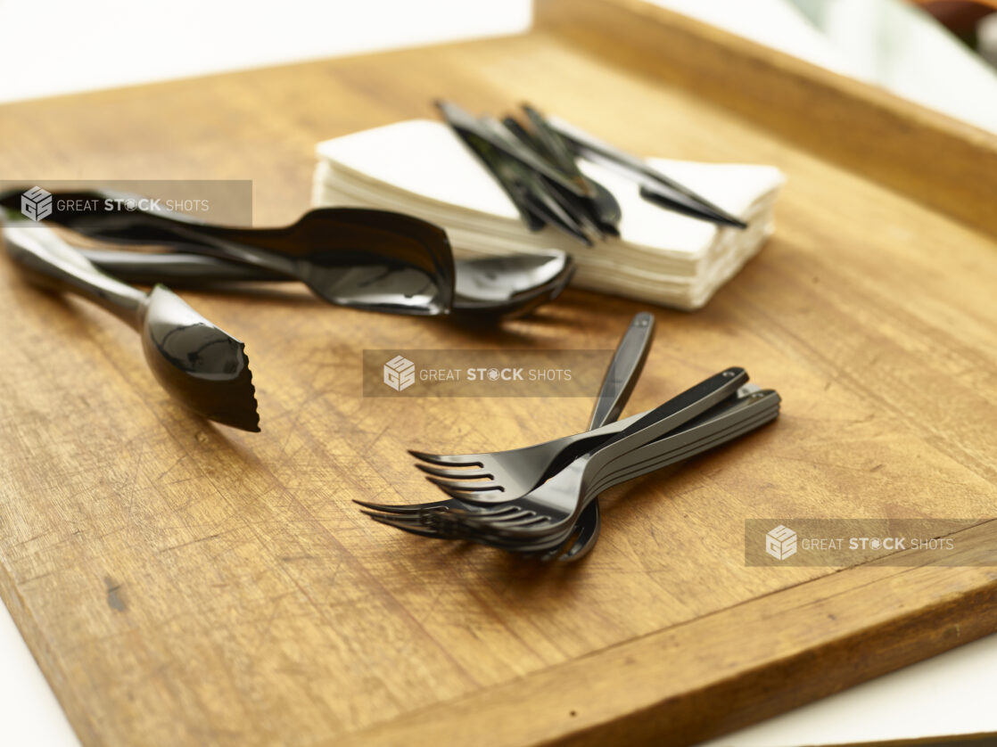 Black disposable plastic cutlery and utensils with paper napkins on a wooden cutting board