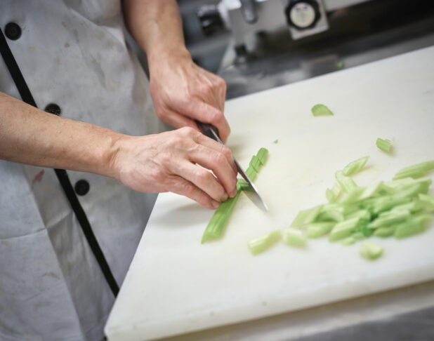 Prep cook chopping celery on a white cutting board, kitchen setting