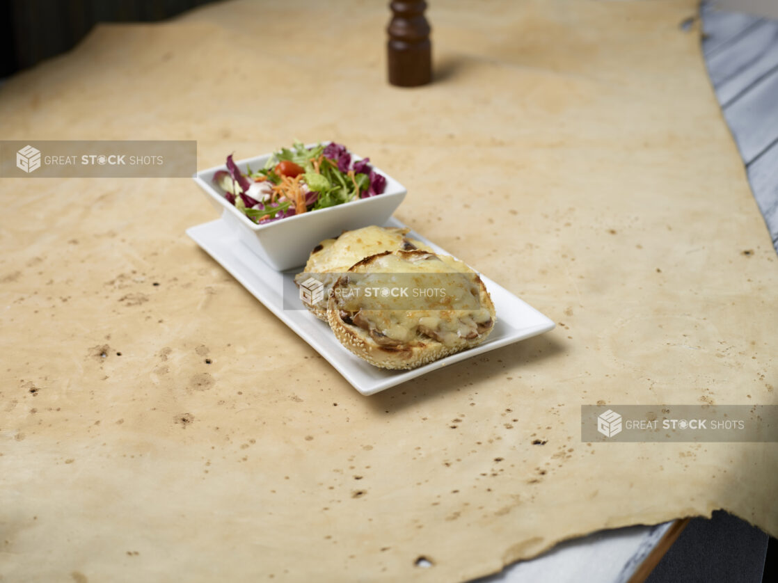 Open-faced toasted sesame seed bagel with mushrooms and melted cheese and a side garden salad on a light background