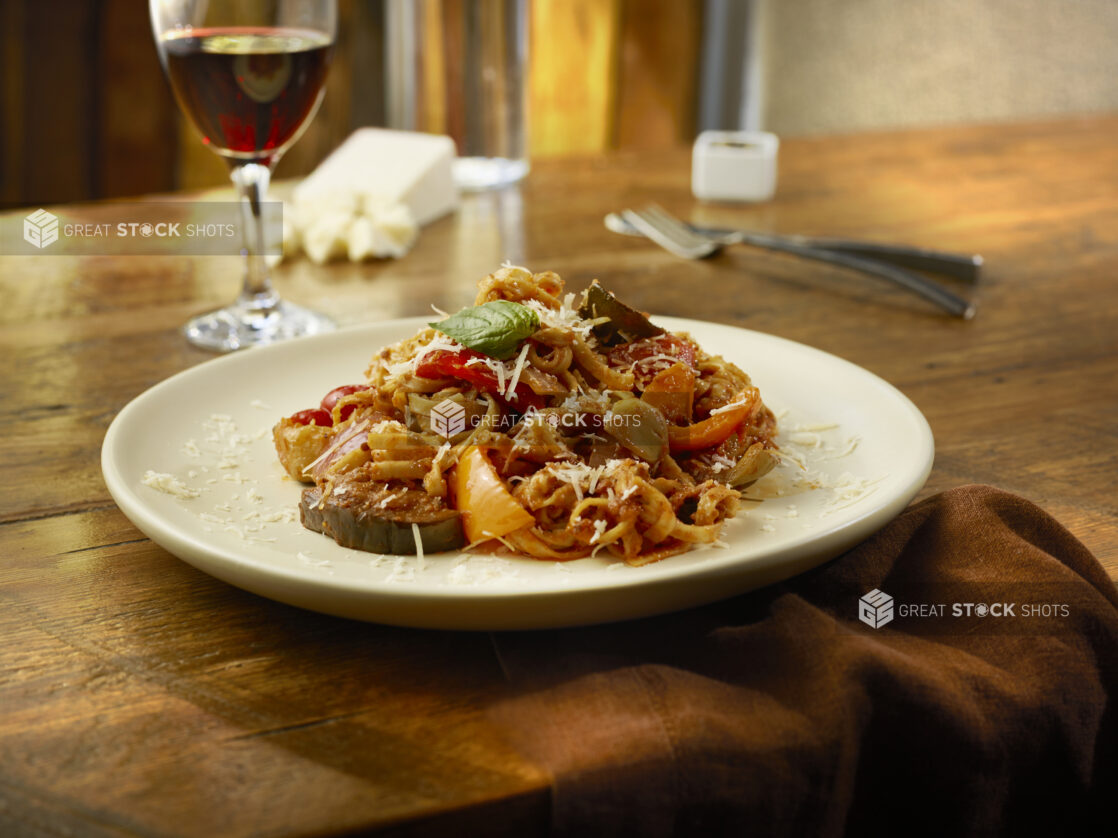 Roasted vegetable fettuccini in tomato sauce in a white bowl on a wooden table with glass of red wine