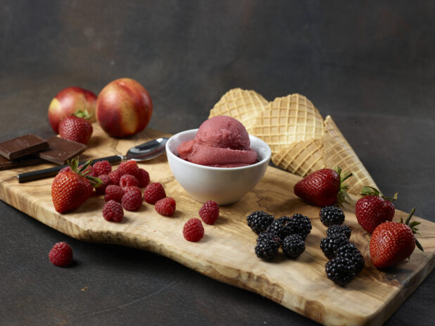 Scoop of red berry gelato in a white bowl surrounded by fruit, chocolate pieces, and waffle cones on a wooden board with a dark background