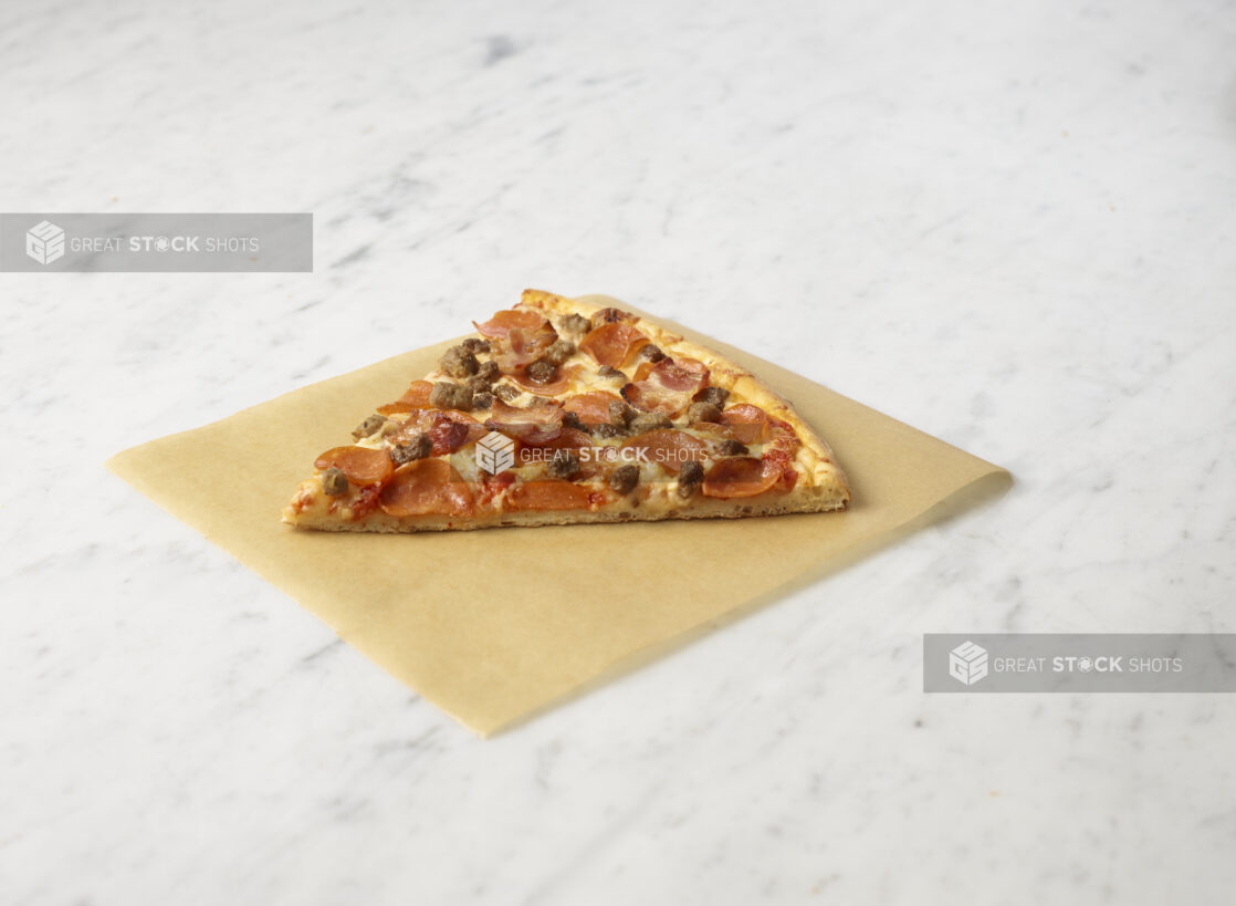 Slice of three meat pizza on parchment paper on a marble background