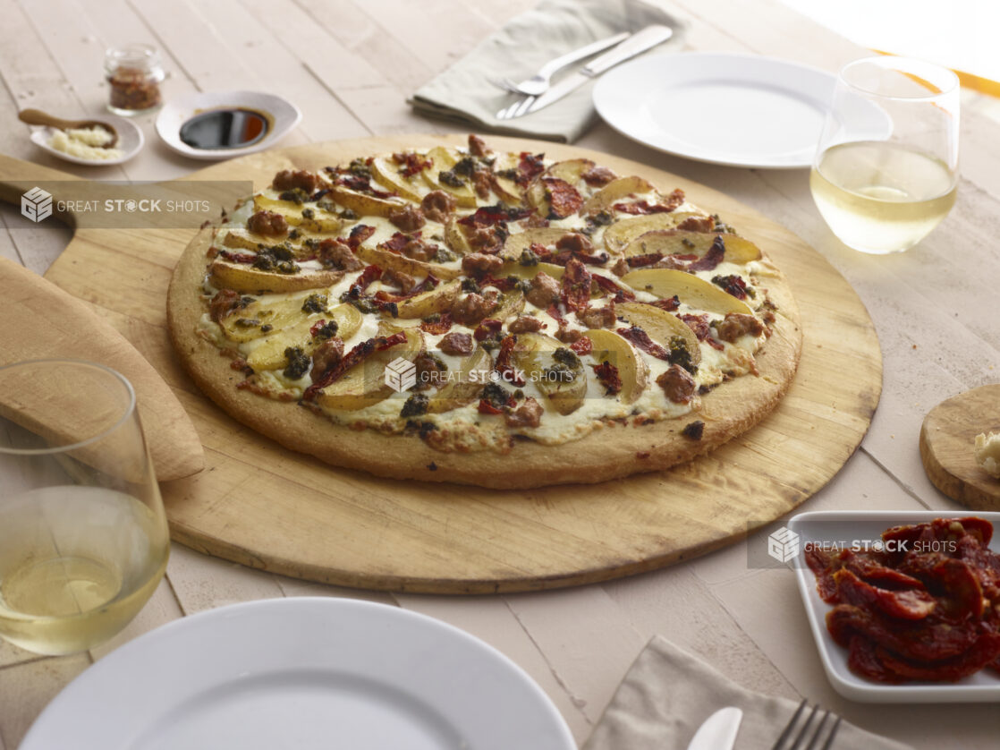 White pizza with potatoes, sun-dried tomatoes and pesto on a paddle, dinner table items surrounding