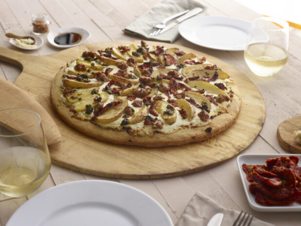 White pizza with potatoes, sun-dried tomatoes and pesto on a paddle, dinner table items surrounding