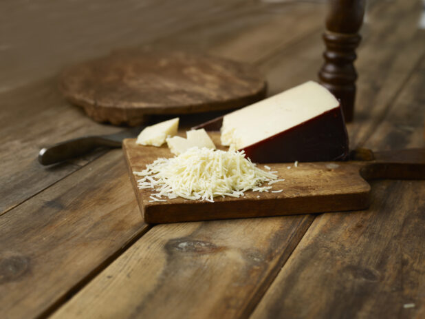 Block of parmesan cheese with shredded pieces and chunks on a small wooden cutting board on a wooden background