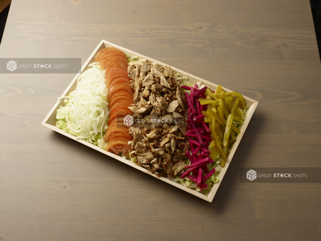 Wooden catering platter with sliced chicken shawarma, tomatoes, onions, pickles and turnip on a bed of lettuce on top of a wooden background