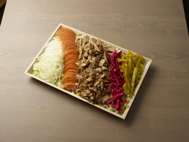 Wooden catering platter with sliced chicken shawarma, tomatoes, onions, pickles and turnip on a bed of lettuce on top of a wooden background