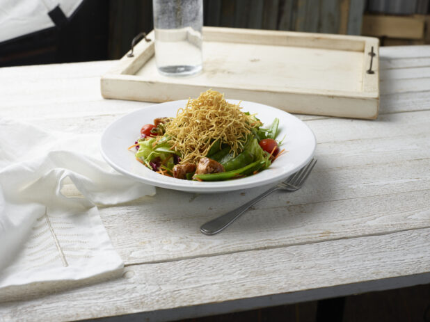 Asian salad with crunchy noodles on top in a white bowl on a whitewashed wood background