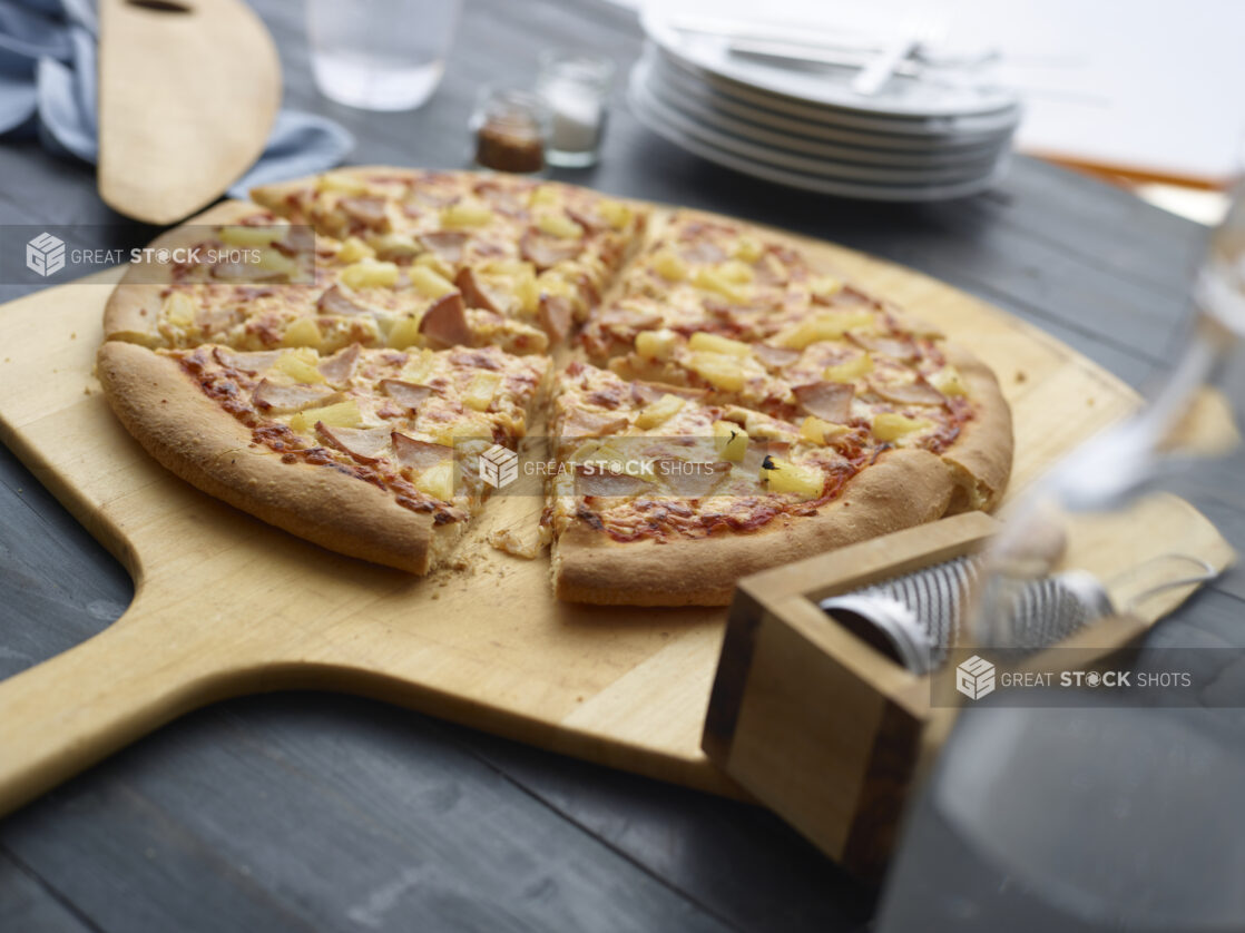 Hawaiian pizza cut into slices on wooden pizza paddle with a dark background