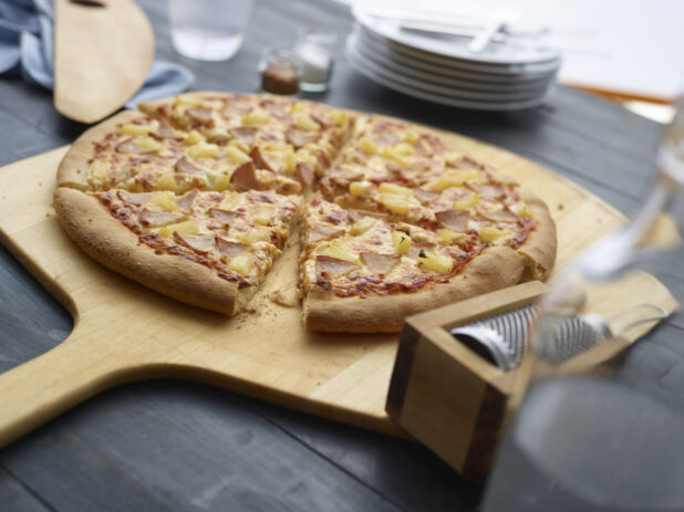 Hawaiian pizza cut into slices on wooden pizza paddle with a dark background