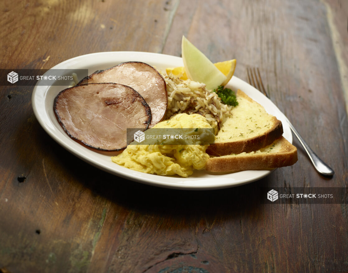 Breakfast plate of scrambled eggs, fried ham, hashbrowns and toast on a wooden background