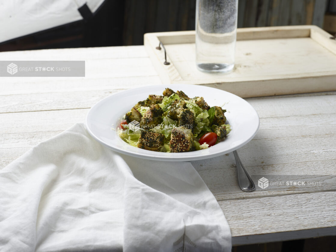 Green salad with croutons in a white bowl on a white washed wood background with napkin