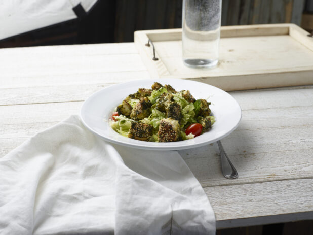 Green salad with croutons in a white bowl on a white washed wood background with napkin