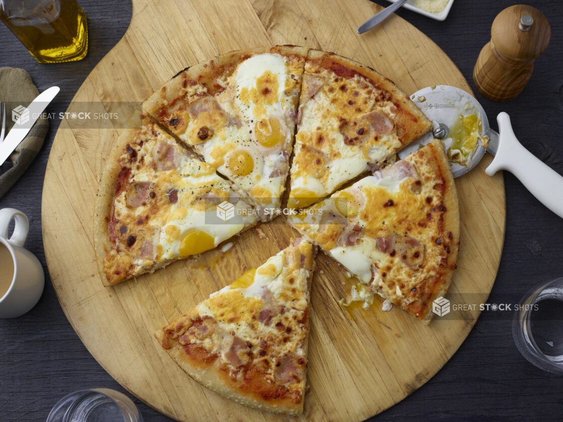 Breakfast egg and ham pizza cut into slices on a wooden cutting board overhead