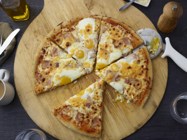 Breakfast egg and ham pizza cut into slices on a wooden cutting board overhead