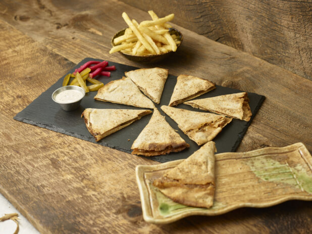 Cut lahm bi ajeen/middle eastern pizza on a slate platter with pickles, pickled turnip, dip, bowl of french fries, side platter with a piece of lahm bi ajeen all on aged wooden table